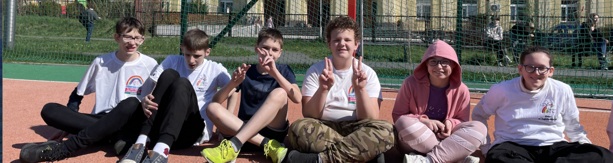 Dzieci siedzą na boisku przed bramką. W tle budynek szkoły.